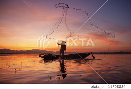 Fisherman casting his net on during sunset. Silhouette Asian fisherman on wooden boat casting a net for freshwater fish. Thailand culture. 117088844