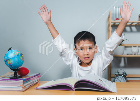 Concept of education and homework and school. Little boy smiling and raising his hand happily while sitting at a table with a book. Children celebrating finishing their homework. 117095375