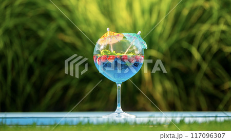 Glass with a blue cocktail, garnished with fruit and mini umbrellas, on the pool edge with green grass in the background 117096307