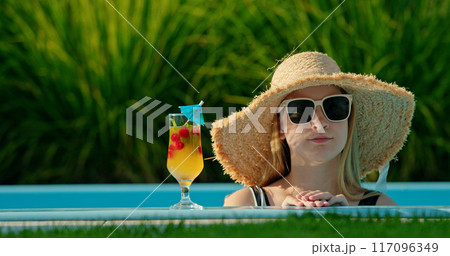 Teenage girl in a hat peeking out of a pool with a cool drink beside her 117096349