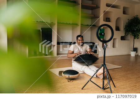 Happy smiling black male blogger practicing native music traditional drumming instrument sitting on floor in front of mobile phone with ring light. Concept of sound therapy, mindfulness, tranquility. Happy smiling black male blogger practicing native music traditional drumming instrument sitting on floor in front of mobile phone with ring light. Concept of sound therapy, mindfulness, tranquility. 117097019