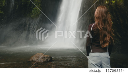Tourist girl standing near powerful jungle waterfall. Long hair blows in wind. Tourism, trekking, hiking, relax, active lifestyle, summer holiday vacation on Bali. Slow back view zoom in 117098334