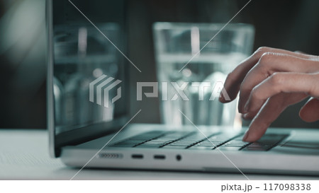 Female hands typing on computer close up. Woman user worker typing on laptop notebook keyboard desk working online with pc software apps. Drink water from glass, keep health balance hydration indoors Female hands typing on computer close up. Woman user worker typing on laptop notebook keyboard desk working online with pc software apps. Drink water from glass, keep health balance hydration indoors 117098338