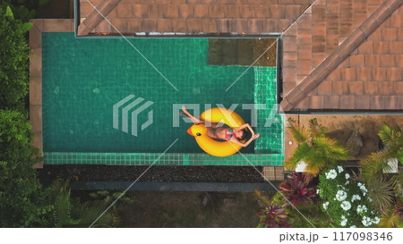 Young woman is enjoying a sunny day, floating on a yellow inflatable duck in a luxurious swimming pool. The image transmits tranquility and the pleasure of a relaxing moment 117098346