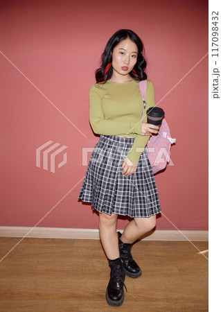 Vertical full shot of confident young Asian female student wearing plaid skirt in preppy style holding reusable coffee cup against pinkish red wall, copy space 117098432