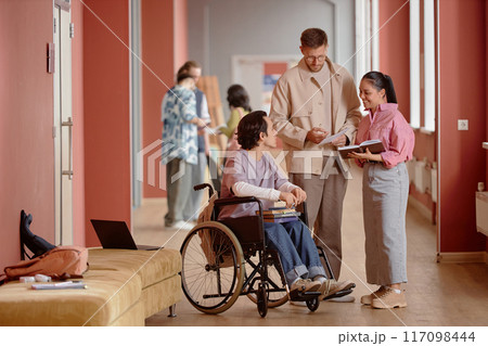 Adult male professor answering question in friendly informal conversation with male student using wheelchair and smiling female learner in pink university hallway 117098444
