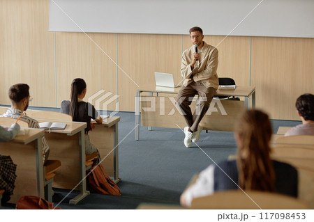 Adult male teacher with hand held microphone lecturing university students while leaning on desk in lecture hall, copy space 117098453