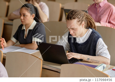 Young male dreadlocked student studying using laptop during lecture class at university. Concentrated young man with tattoo on arm writing on computer 117098456