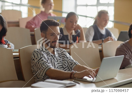 Yong Middle Eastern male student with neat beard studying using laptop in lecture class wearing glasses at university, copy space 117098457