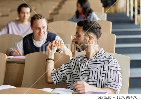 Side view shot of young male Middle Eastern student at desk secretly passing classmate behind him note while attending class in lecture hall at university 117098470