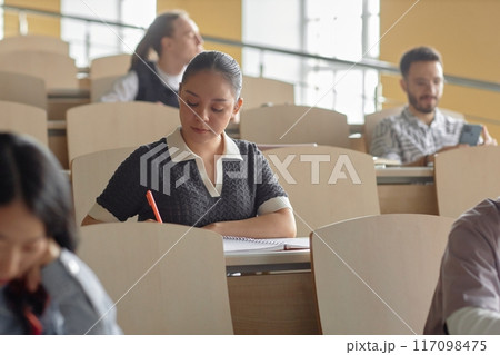 Portrait shot of concentrated young Hispanic female student note taking while sitting at desk in university lecture hall, copy space 117098475