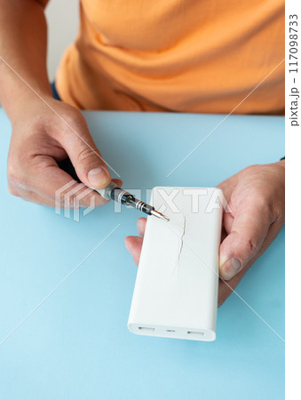 Close up of hands repairing white power bank with screwdriver on blue background. Close up of hands repairing white power bank with screwdriver on blue background. 117098733