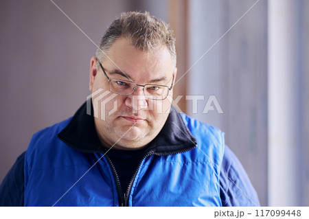 Large, plump-cheeked man of Caucasian ethnic group in prescription glasses has frowning look from under his eyebrows. Large, plump-cheeked man of Caucasian ethnic group in prescription glasses has frowning look from under his eyebrows. 117099448