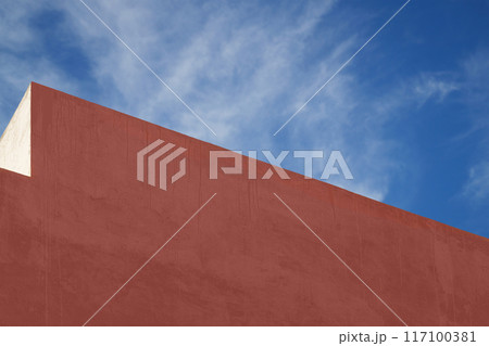 Painted dark red, flat surface of exterior wall of high-rise building without windows with traces of drips against background of blue sky with fuzzy clouds. Blank for design, text with copy space 117100381