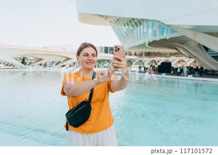Young traveling woman taking selfie outdoors. Concept of travel, tourism and vacation in city. Ciudad de las Artes y las Ciencias de Valencia.Valencia City of Arts and Sciences. High quality photo 117101572