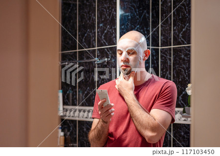 View from door at young adult Caucasian bald bearded man using cloth mask to moisturize the skin of his face and holds tube of cream. Beauty routine and skin care View from door at young adult Caucasian bald bearded man using cloth mask to moisturize the skin of his face and holds tube of cream. Beauty routine and skin care 117103450