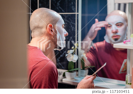 View from door at young adult Caucasian bald bearded man using cloth mask to moisturize the skin of his face. Beauty routine and skin care 117103451