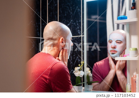 Back view from door of young adult Caucasian bald bearded man using cloth mask to moisturize the skin of his face. Beauty routine and skin care concept 117103452