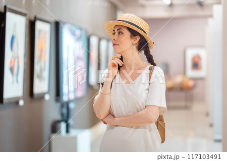 Historic education. Pretty young Caucasian woman wearing straw hat admiring paintings in the gallery. Concept of visit museum and cultural education 117103491
