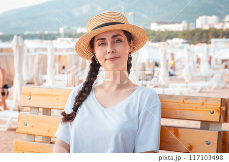 Summer vacation. Portrait of beautiful Caucasian young woman in straw hat sitting on bench at city beach. Concept of travel season and psychology 117103498