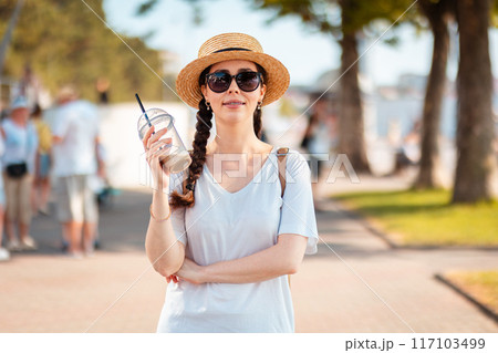 Portrait of young beautiful elegant Caucasian woman wearing straw hat and sunglasses posing at the city boulevard holding drink in her hand. Blurred background. Concept of summer season vacation Portrait of young beautiful elegant Caucasian woman wearing straw hat and sunglasses posing at the city boulevard holding drink in her hand. Blurred background. Concept of summer season vacation 117103499