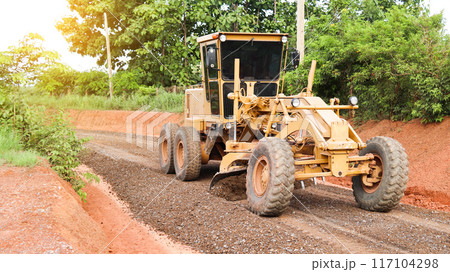 Image of a grader leveling gravels on a dirt road in preparation for asphalt paving for new road construction. 117104298