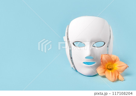 Cosmetic LED facial mask regenerative treatment on blue background with flowers lilies. Health and beauty of the face. 117108204