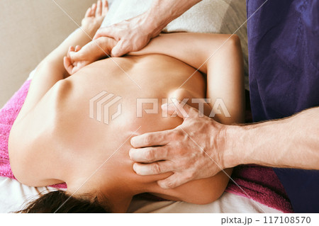 Shoulder area, upper back and neck deep stroking and pressure tissue massage, young white woman receiving physiotherapy while lying face down on massage table. 117108570