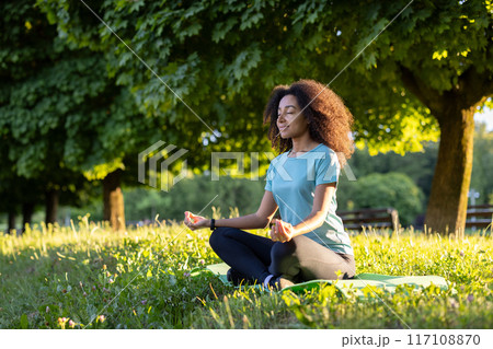 Young African American woman meditating outdoors sitting on grass in yoga pose in park setting. Tranquil moment focusing on calm and mindfulness surrounded by nature during sunny day. 117108870