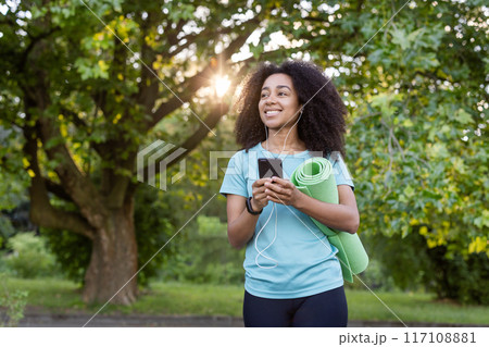 Young African American athlete carrying yoga mat, listening to music, smiling while using smartphone outdoors in park. Fitness enthusiast enjoying nature, technology, and healthy lifestyle. 117108881