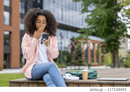 Young woman with curly hair and glasses looks shocked at her smartphone while sitting on a bench in an urban area. Backpack, coffee cup, and laptop next to her. 117108945
