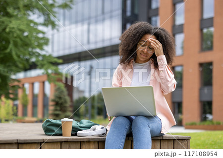 Stressed african american woman student working on laptop on campus. She appears frustrated and exhausted, sitting outside university building with coffee and backpack, showing fatigue with studies. Stressed african american woman student working on laptop on campus. She appears frustrated and exhausted, sitting outside university building with coffee and backpack, showing fatigue with studies. 117108954