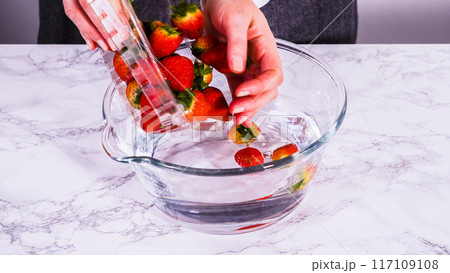 Ripe strawberries are submerged in water within a large glass mixing bowl, a step in washing the fruit to ensure cleanliness and longevity before storage or consumption. 117109108