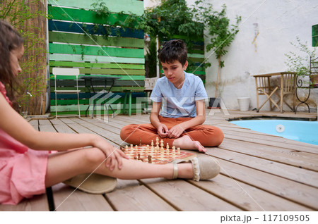 Children playing chess on a wooden deck near the pool in a backyard setting Children playing chess on a wooden deck near the pool in a backyard setting 117109505