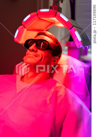Handsome male with hairloss problem in protective goggles having LED light hair photodynamic therapy treatment after hair transplant at clinic. Man client having non-invasive type of phototherapy. 117109890