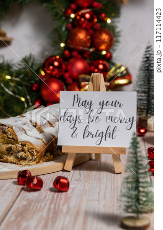 Note with saying MAY YOUR DAYS BE MERRY AND BRIGHT congratulations. Traditional German Christmas stollen cake in background of handmade festive decor. Winter holiday treats. Vegan baking recipe Note with saying MAY YOUR DAYS BE MERRY AND BRIGHT congratulations. Traditional German Christmas stollen cake in background of handmade festive decor. Winter holiday treats. Vegan baking recipe 117114423