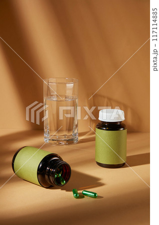 Brown background photography with medicine concept for advertisement from front view. Two brown supplement bottles with blank green label, featured on the table next to a glass of water 117114885