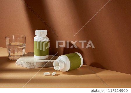 The empty green label pill bottle displayed on yellow countertop, over brown background with some scattered white pills and a glass of water. High angle shot photo, blank space for designing 117114900