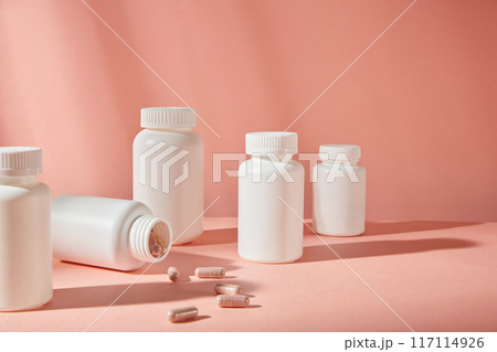 Several white medicine bottle without label standing out on pink background, some purple capsules scattered in front. Copy space for adding text, frontal shot Several white medicine bottle without label standing out on pink background, some purple capsules scattered in front. Copy space for adding text, frontal shot 117114926