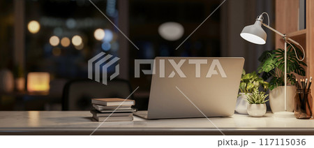 A laptop computer on a table in a dark room at night, illuminated by a table lamp. 117115036