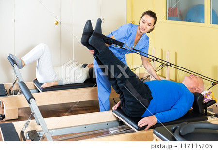 Rehabilitation physician controlling senior man exercising on pilates reformer 117115205