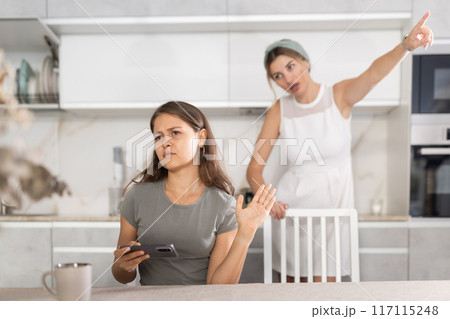 Upset daughter is sitting at table. Mom yells at her daughter in background Upset daughter is sitting at table. Mom yells at her daughter in background 117115248