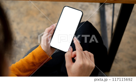 A woman sitting indoors and using her smartphone. a smartphone with a white-screen mockup. 117115692