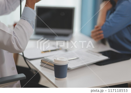 Doctor and patient sitting near each other at the wooden desk in clinic. Female physician's pointing to a records form. Medicine concept 117118553