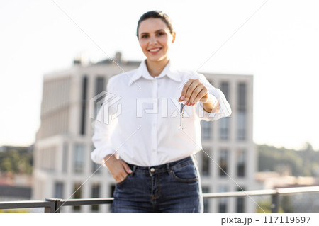 Smiling woman holding keys with modern building in background Smiling woman holding keys with modern building in background 117119697