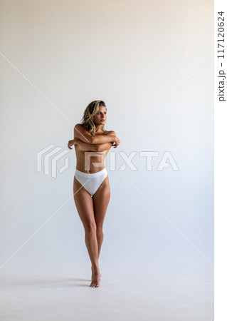 Beautiful blonde posing on a white background in a photo studio in white panties. Covers her breasts with her hands. Full-length photo 117120624