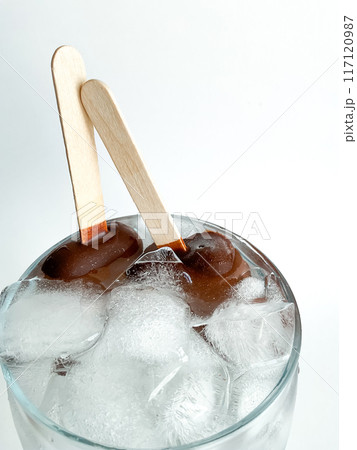 Two popsicle chocolate covered ice cream bars on wooden sticks placed in glass bowl with ice cubes. Top view of frozen treats with condensation on white background. 117120987