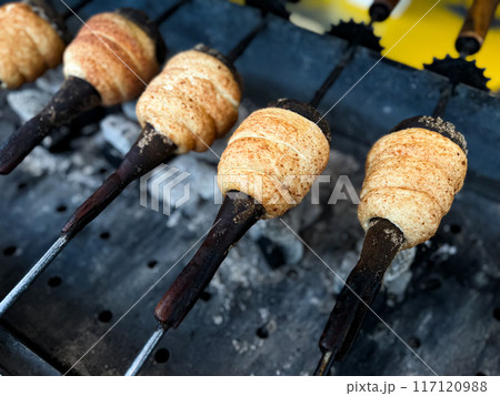 Trdelnik baking on metal rods over hot coals. Close up view of traditional Hungarian street food being prepared at market. Food and culinary concept for poster and print 117120988