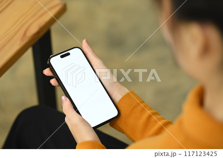 View over shoulder of young woman in yellow sweater holding smartphone with blank screen 117123425