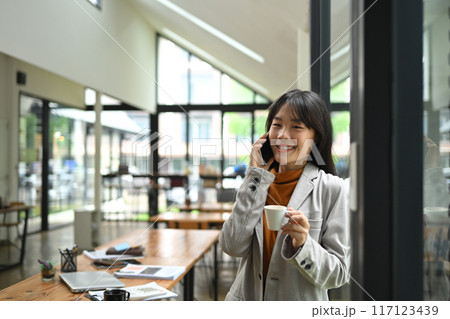 Charming young businesswoman having pleased phone conversation while drinking coffee by window 117123439